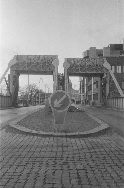 A river with a concrete bridge spanning across it, supporting vehicular traffic. A house removal process is visible with a moving van parked on the pavement and several cardboard boxes, wrapped furnit