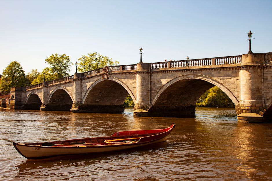 A historic stone bridge with multiple arches spanning a river, featuring decorative lampposts along its length and a balustrade on top. Below the bridge, the water reflects the warm sunlight, creating