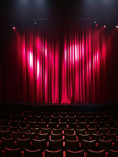 Inside the auditorium of the Rose Theatre, there is a large stage with deep red velvet curtains drawn closed, illuminated by soft lighting that highlights the fabric's texture and folds. In front of t